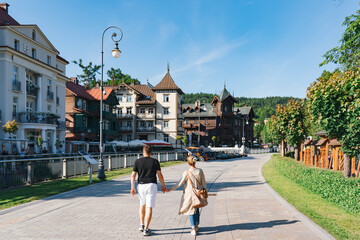 A couple strolls hand in hand along a sunlit promenade in Krynica, resort town in southern Poland, framed by historic villas, leafy trees, and forested hills under a clear blue sky.
