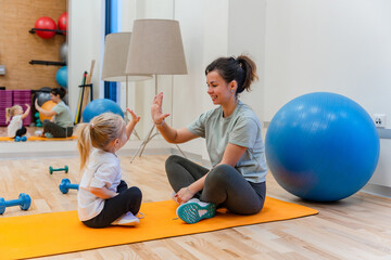 Mother and daughter child sit on the mat and high-five each other in the gym