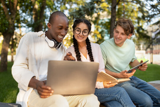 Three friends gather in a park on a sunny day, focused on a laptop. They appear engaged and happy, sharing ideas and collaborating in a relaxed setting.