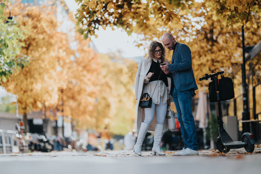 A stylish couple stands on a autumn city street, looking at a phone with a scooter nearby. Warm fall colors, casual chic outfits, and a relaxed urban mood.