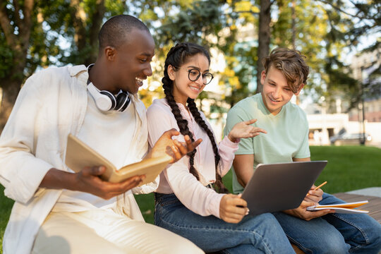 Three friends enjoy a sunny day while studying in a park. They are engaged in discussion, using a laptop and taking notes, surrounded by trees and greenery.