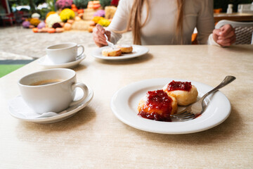 Cottage cheese pancakes with jam served with coffee at cafe table, morning meal composition with female figure in soft background, warm food atmosphere
