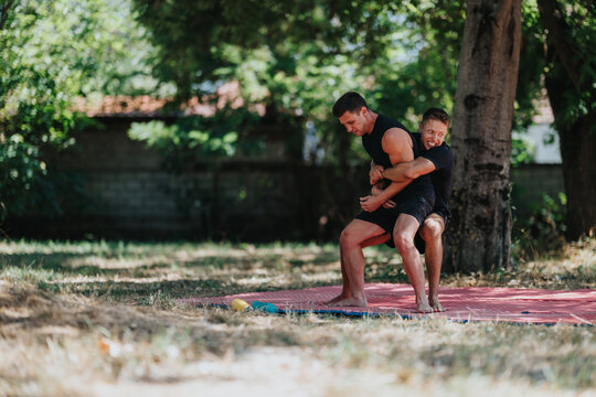 Two men wrestle on a red mat in a sunlit park, displaying strength, teamwork, and competitive fitness. Ideal for sports, outdoor activity, and athletic training themes. - Powered by Adobe