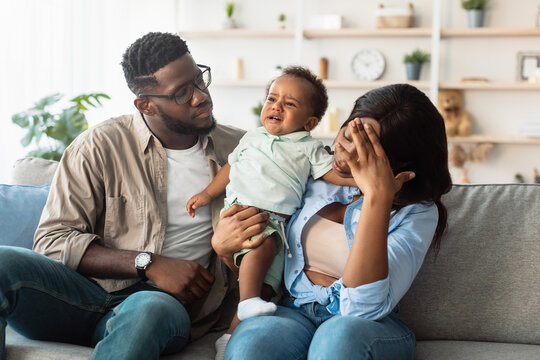Two tired African American parents sit on a comfortable sofa in their living room. The father holds their crying baby while the mother rubs her head in frustration.