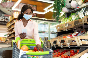 In a bustling grocery store, a woman wearing a mask selects fresh fruits and vegetables for her cart while using her smartphone for shopping assistance and price checks.