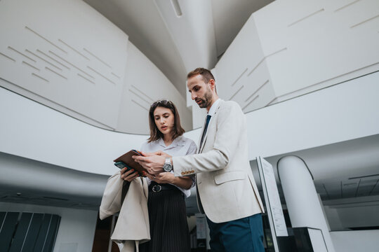 A man and a woman in business attire stand in a sleek, contemporary lobby, examining a tablet together. The scene conveys teamwork, planning, and professional collaboration in a corporate setting.