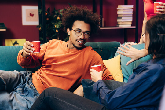 Young friends relaxing on sofa, drinking beverages, and enjoying a home party