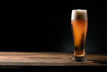 Cold tall glass of amber beer with foam on a wooden table against a dark background, showing condensation and warm light, idea for bar menus, brewery promotions, and drink visuals
