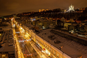 Aerial drone view of a snowy city street with Christmas lights at night.