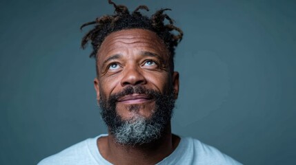 A mature man with dreadlocks and a salt-and-pepper beard gazes upwards, embodying contemplation and hope, set against a soft gray background that emphasizes his expression.