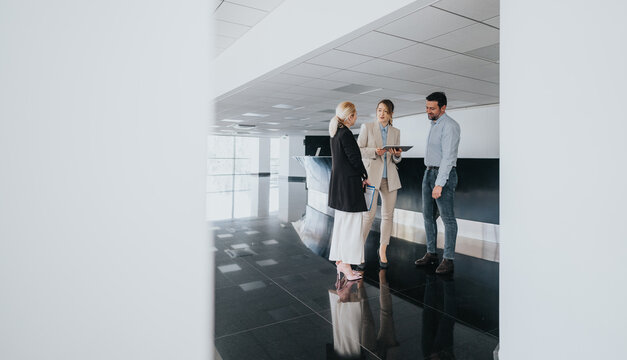 Three coworkers stand in a bright, modern office lobby, discussing a tablet and planning together. The polished, reflective floor adds a sense of professionalism and collaborative energy.