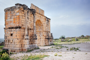 Fototapeta premium Ancient stone archway ruins stand majestically against a cloudy sky, surrounded by grassy terrain, showcasing historical architecture and remnants of a bygone era