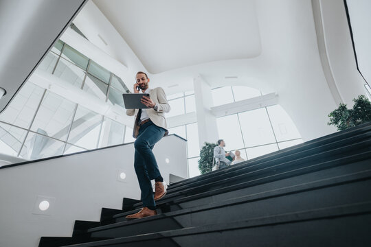 A confident businessperson navigates a sleek, sunlit atrium, using a tablet while on a call. In the background, people chat near large windows, suggesting collaboration and a dynamic work environment.