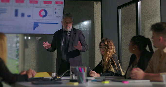 Corporate manager explaining report data to attentive colleagues in boardroom with screen displaying colorful charts and graphs - Powered by Adobe