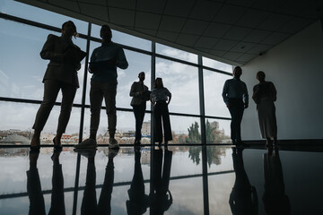 A group of colleagues stands in shadow along a glass wall, their silhouettes reflecting on the polished floor. The cityscape behind them hints at a collaborative business meeting in progress.