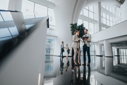 Two colleagues in smart suits talk and review a tablet in a bright, contemporary office lobby as others mingle in the background.