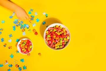 A vibrant top-down view of colorful popcorn in two bowls, scattered puzzle pieces, and a child’s hand reaching out, all set against a bright yellow background.