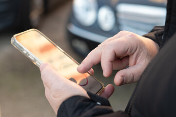 Close-up of a person using a smartphone to pay for parking through a mobile app, standing near a parked car and tapping on the screen.