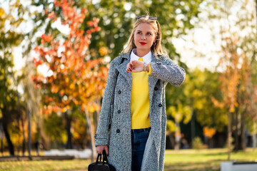 Urban businesswoman with sunglasses in stylish autumn coat checking the time on her wristwatch while walking with a laptop bag through the city park during sunny day.