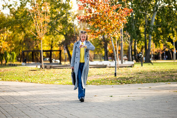 Confident businesswoman with sunglasses in stylish autumn coat checking the time on her wristwatch while walking with a laptop bag through the city park during sunny day.