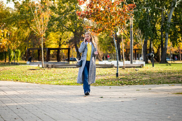 Modern businesswoman in autumn coat talking on the smartphone while walking through city park on a sunny day.