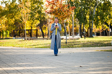 Urban businesswoman in warm stylish coat using smartphone while walking through park in the city during sunny autumn day. 