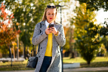 Fashionable businesswoman with sunglasses in modern autumn coat using smartphone while walking  through the city park during sunny day.