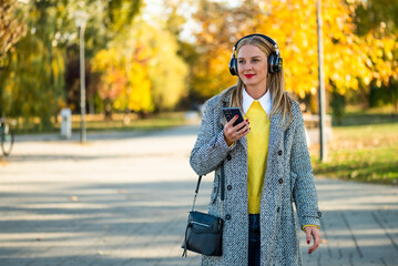Modern businesswoman in stylish coat using wireless headphones and smartphone while walking through the city park during sunny autumn day.