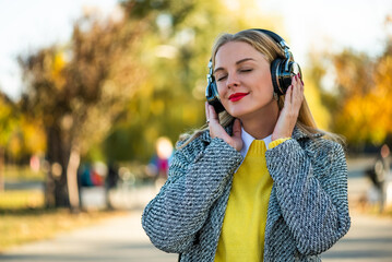 Close up portrait of beautiful urban businesswoman in autumn coat listening to music through wireless headphones while standing in the city park during sunny autumn day.