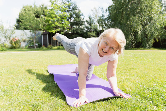 Active senior woman practicing yoga on mat in garden, smiling while exercising outdoors on a sunny day