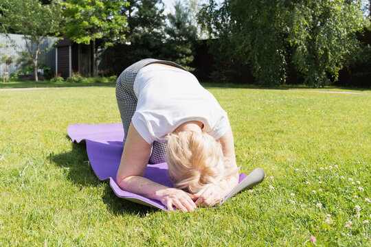 Elderly woman in forward bend yoga pose on mat in backyard, focusing on flexibility and relaxation outdoors