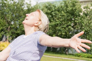 Obraz premium Senior woman smiling with eyes closed and arms wide open, enjoying sunshine and freedom in a garden
