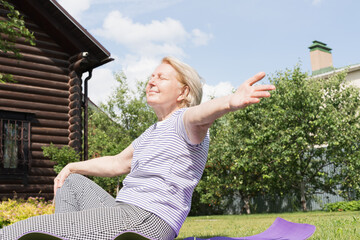 Elderly woman sitting on grass with arms wide open, smiling and enjoying sunshine in a peaceful backyard