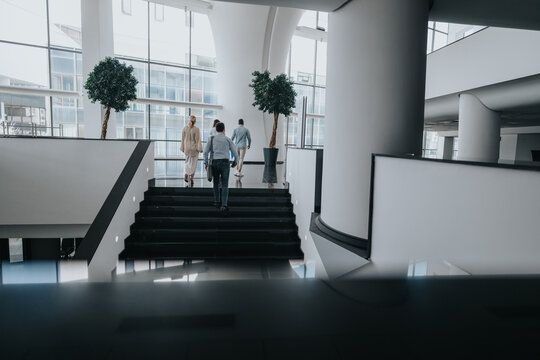 A modern office atrium with glass walls and large columns. Several people walk up a black staircase, carrying bags and folders, creating a sense of travel, collaboration and daily work in a workplace.