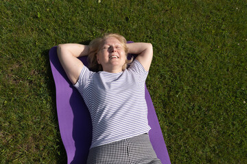 Senior woman relaxing on a yoga mat outdoors, lying on the grass with closed eyes and enjoying the sunlight. Peaceful moment of rest, wellness, and mindful outdoor lifestyle.