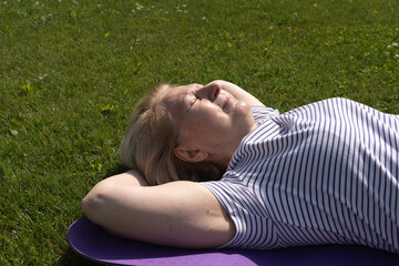 Senior woman lying on a yoga mat outdoors, relaxing on the grass with eyes closed and enjoying the warm sunlight. Peaceful moment of rest, wellness, and outdoor lifestyle.