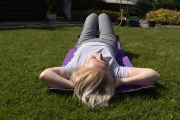 Elderly woman lying on a mat in the garden, relaxing with hands behind head on a sunny summer day