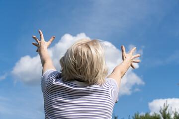 Senior woman raising arms toward the sky, enjoying a sunny day with blue sky and clouds. Seems like she is holding white cloud