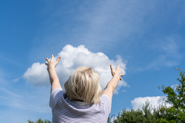 Senior woman raising arms toward the sky, enjoying a sunny day with blue sky and clouds. Seems like she is holding white cloud