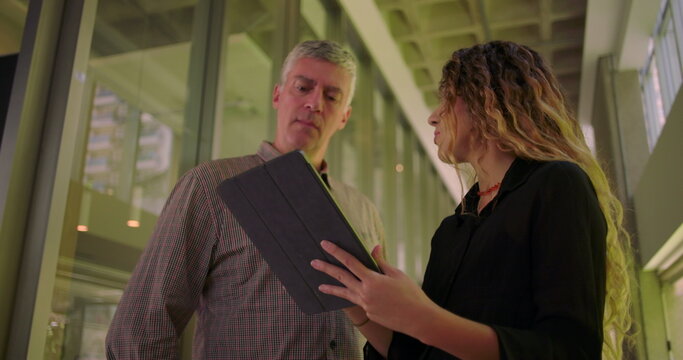 Coworkers standing together in hallway, reviewing information on tablet, collaborative exchange in professional environment, man listening attentively