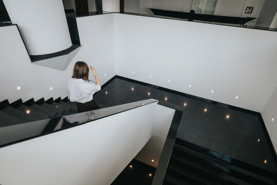 A woman on a black staircase in a contemporary office building, gazing upward. White walls, warm lights, and minimalist design create a calm, aspirational mood in a modern business space.