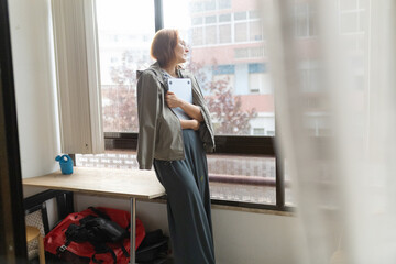 Mature woman holding laptop looking out window contemplating
