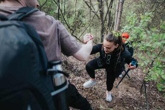 Two hikers on a wooded trail assist each other as they ascend a rocky, steep path. Backpacks, trekking poles, and athletic footwear highlight teamwork, fitness, and outdoor adventure in nature. - Powered by Adobe