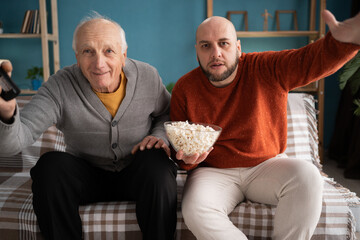 Family leisure time: happy middle aged son and senior father watching exciting football match on large television screen at home, cheering and eating popcorn.
