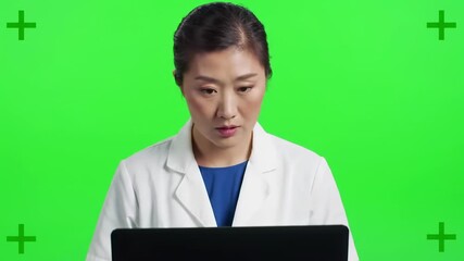 A focused Asian woman in a lab coat looks at a laptop on a chroma-key green screen background. Four green markers are in corners