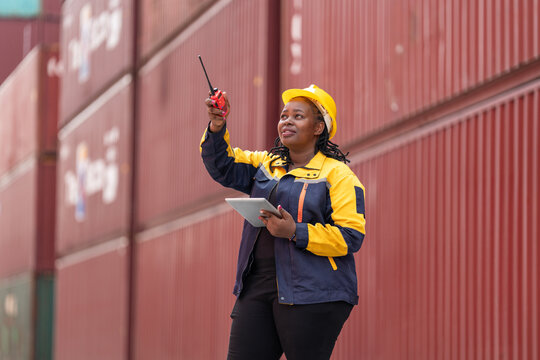 Confident female worker in safety gear uses walkie-talkie and tablet while overseeing shipping containers in logistics yard - Powered by Adobe