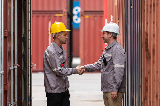 Male warehouse workers in safety helmets shake hands between cargo containers. agreement, teamwork, or successful negotiation.