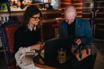 A man and a woman sit at a cafe table, reviewing documents and a laptop. They appear focused and engaged, suggesting a collaborative business discussion and planning session.