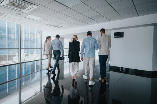 A group of colleagues stroll along a sunlit, glass-walled corridor. They appear to be heading to a meeting, showcasing teamwork, a contemporary workplace, and smart attire in a corporate setting.
