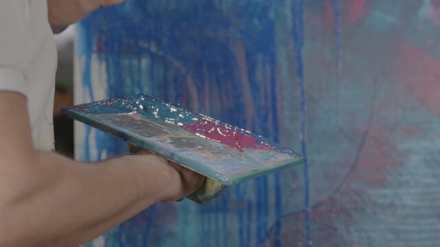 Hand h&auml;lt farbbedeckte Plexiglasplatte vor bemalter Leinwand &ndash; k&uuml;nstlerische Studioszene mit nasser Farbe, Acrylstruktur und kreativem Malprozess